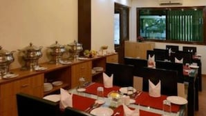 A dining area with tables set for service, a buffet line with food and drink containers, and a window with blinds. - Kastor International (New Delhi)