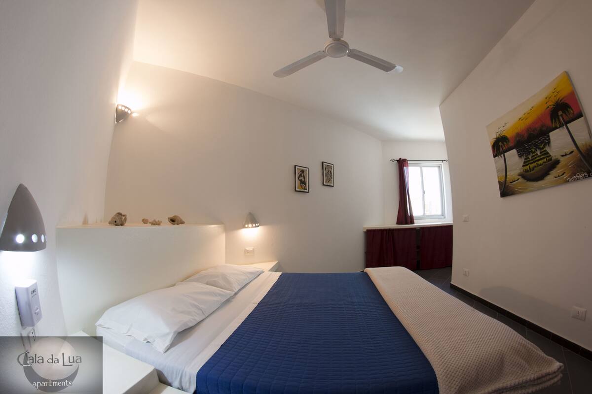 Ein Schlafzimmer mit Bett, Deckenventilator, Nachttisch, Wandlampe und einem Bild an der Wand.