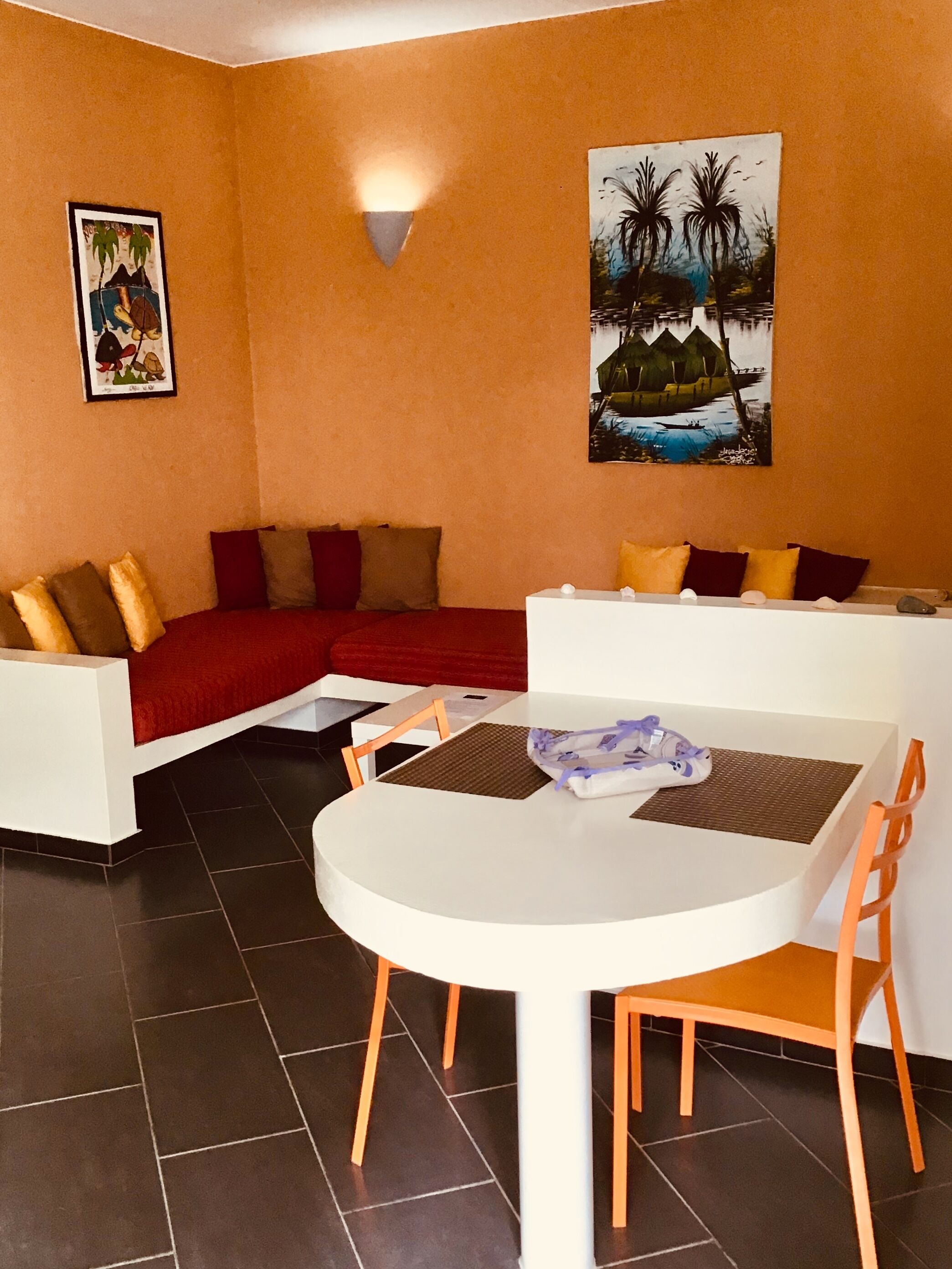 A dining area with a white table and orange chairs, a red sofa with multiple cushions, and two framed pictures on the walls.