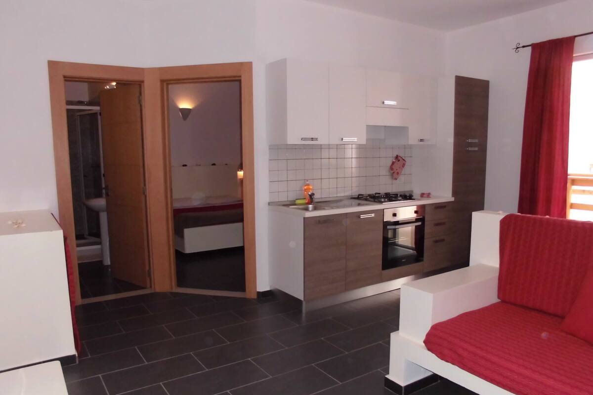 Eine kleine Küche mit Herd, Backofen und weißen Schränken. Ein rotes Sofa und ein kleiner Esstisch sind vorhanden. Durch eine offene Tür ist ein Badezimmer zu sehen.