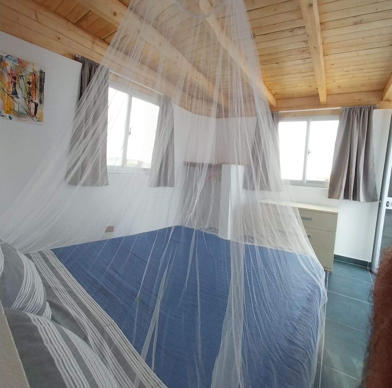 Un dormitorio con cama cubierta por una mosquitera, vigas de madera en el techo, una ventana con cortinas y una pared con un cuadro.
