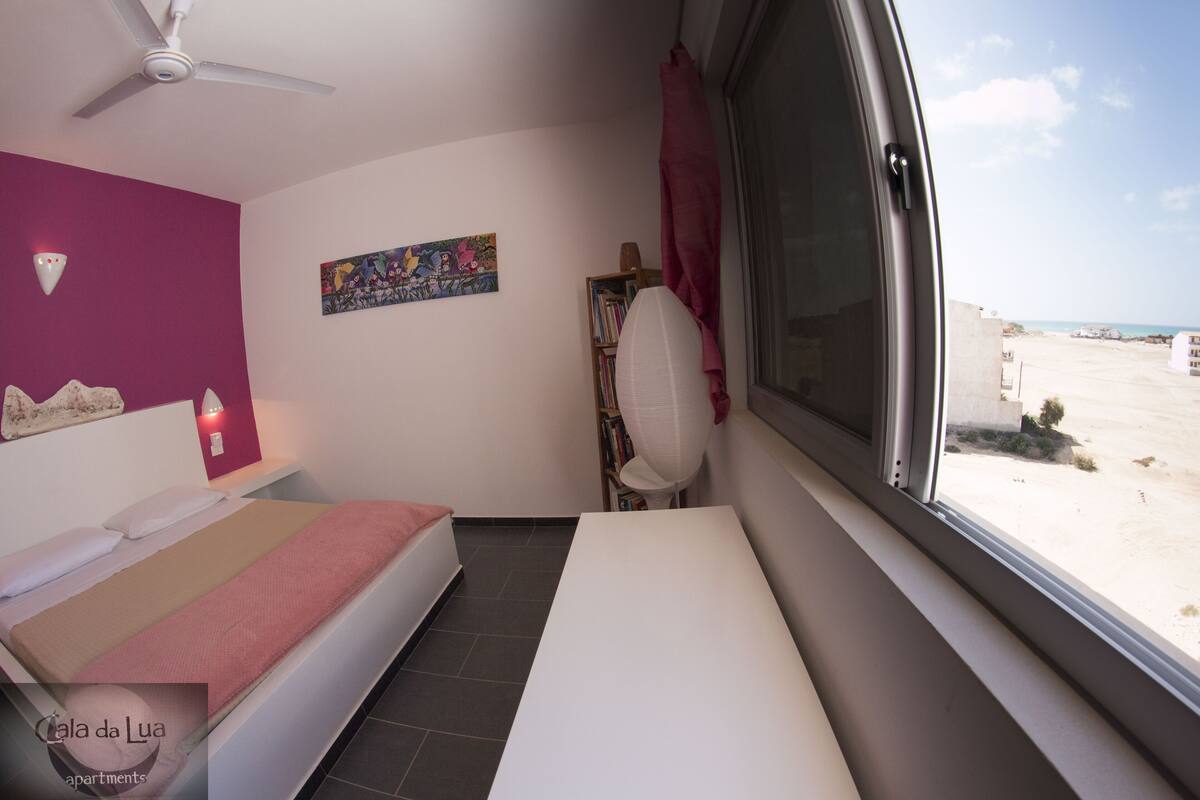 Ein Schlafzimmer mit einem Bett, einem Deckenventilator, einem Fenster mit Blick auf den Strand und einem Bücherregal.