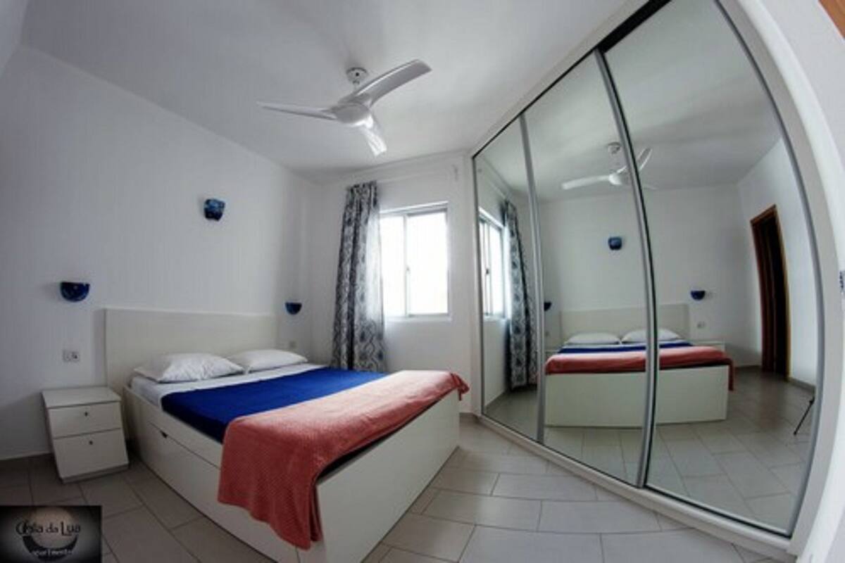 Ein Schlafzimmer mit Bett, Nachttisch, Deckenventilator, Fenster mit Vorhängen und einer Glasschiebetür, die in einen anderen Raum führt.