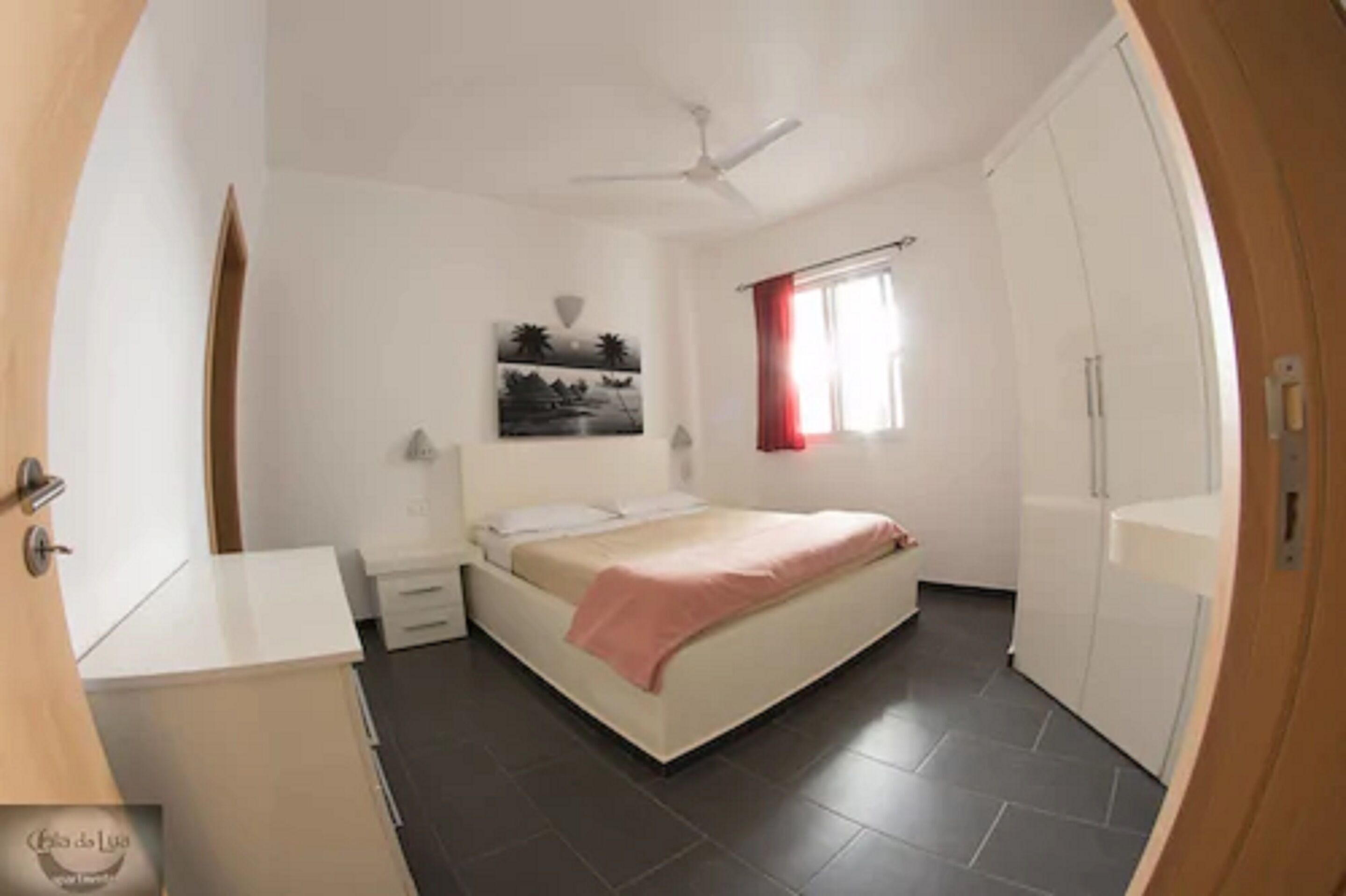 Un dormitorio con cama, mesitas de noche, ventilador de techo, ventana con cortinas rojas y armario.