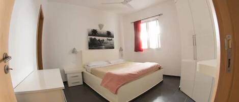 Un dormitorio con cama, mesitas de noche, ventilador de techo, ventana con cortinas rojas y armario.