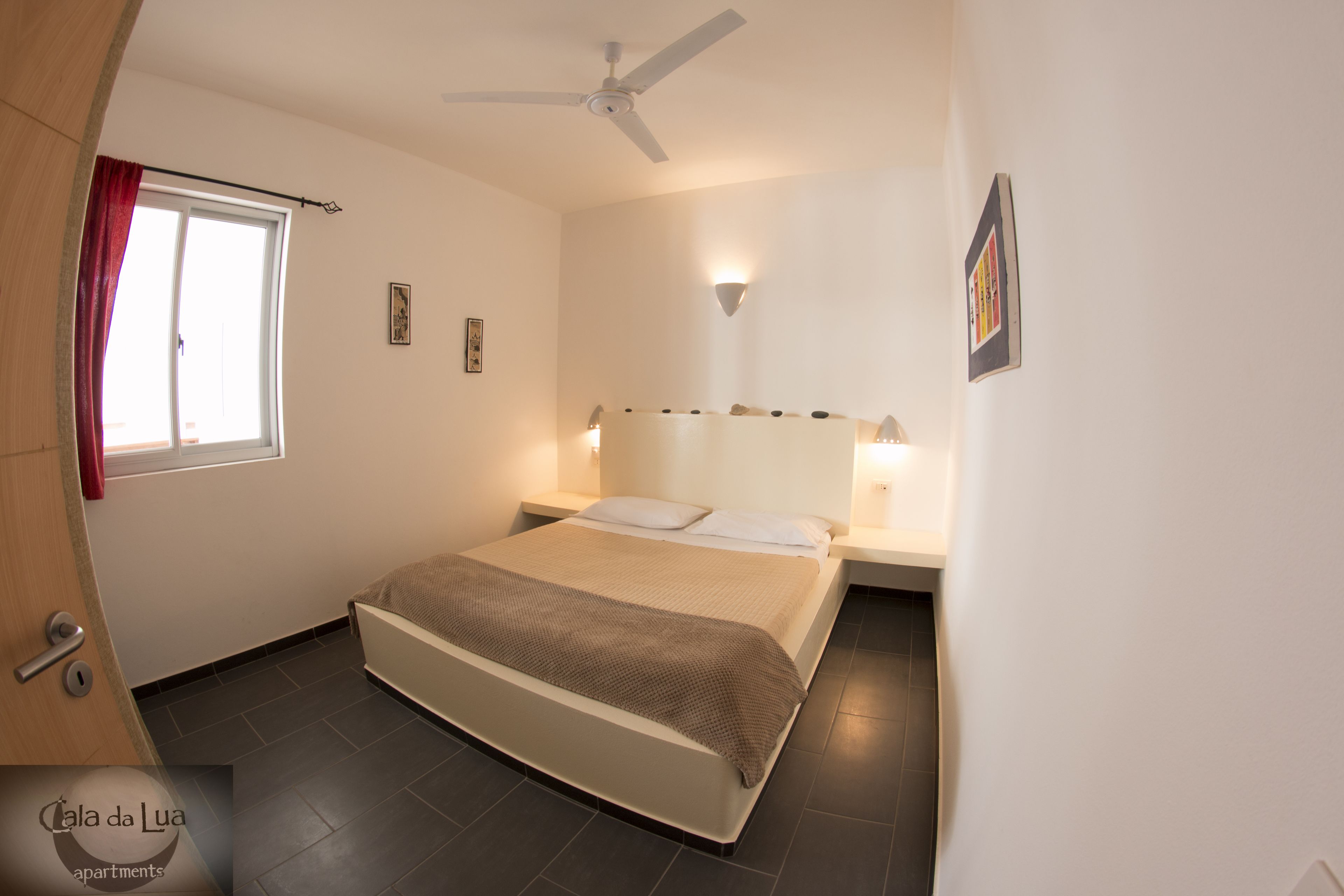 Une chambre à coucher comprenant un lit, un ventilateur de plafond, une fenêtre avec des rideaux, une table de chevet avec une lampe et un tableau accroché au mur.