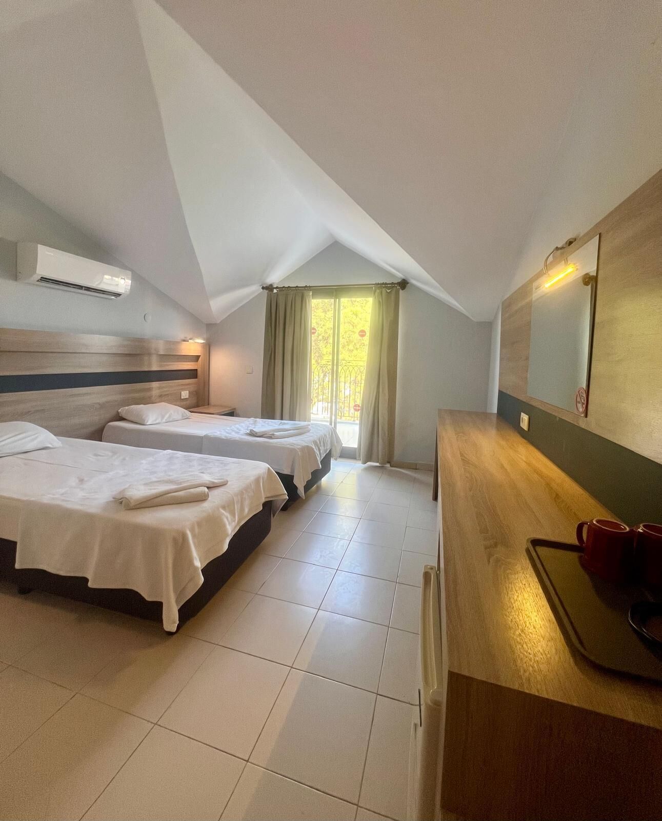 A hotel room with two beds, a wooden desk, a mirror, and a window with curtains.