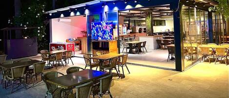 An outdoor dining area with tables and chairs, a glass enclosure, and a television screen displaying a show.