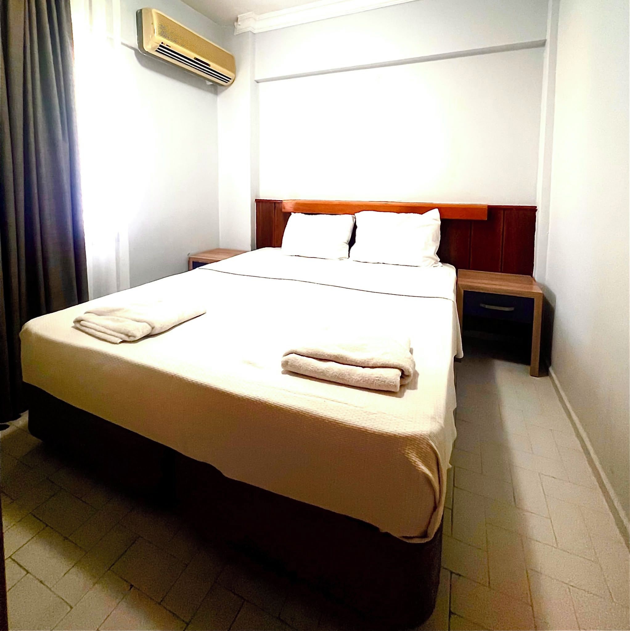 Een hotelkamer met een bed, nachtkastje, wandairconditioner en een raam met gordijnen.
