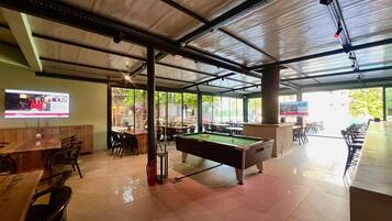 A billiards table in a well-lit indoor area with tables and chairs, a fire extinguisher, and a television screen on the wall.