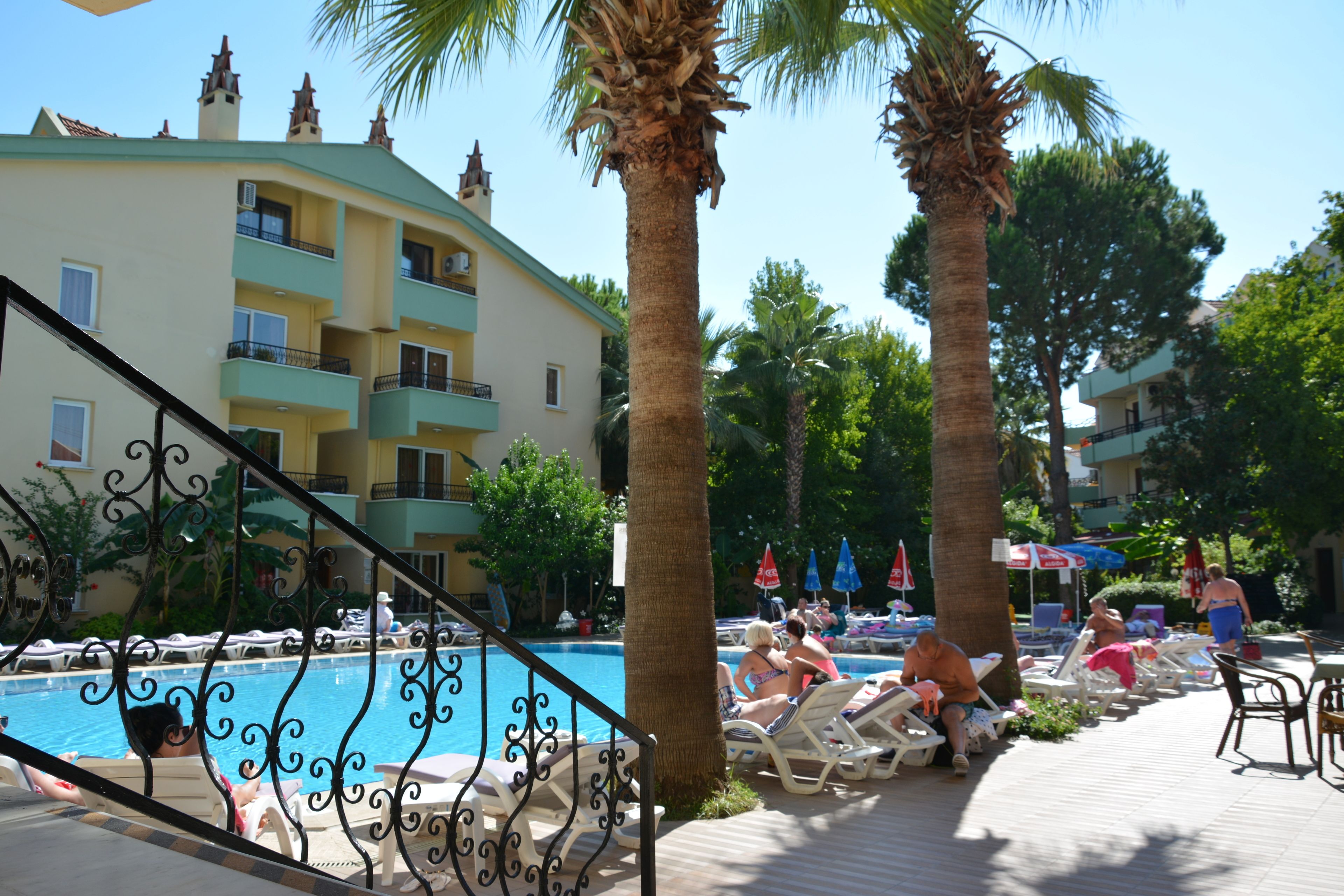 A poolside area with lounge chairs and umbrellas, a palm tree, and a building with balconies.