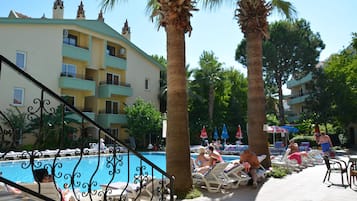 A poolside area with lounge chairs and umbrellas, a palm tree, and a building with balconies.