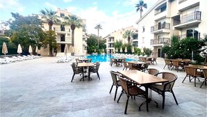 Een zwembad van een resort met ligbedden en tafels, omgeven door palmbomen en een helderblauwe lucht.