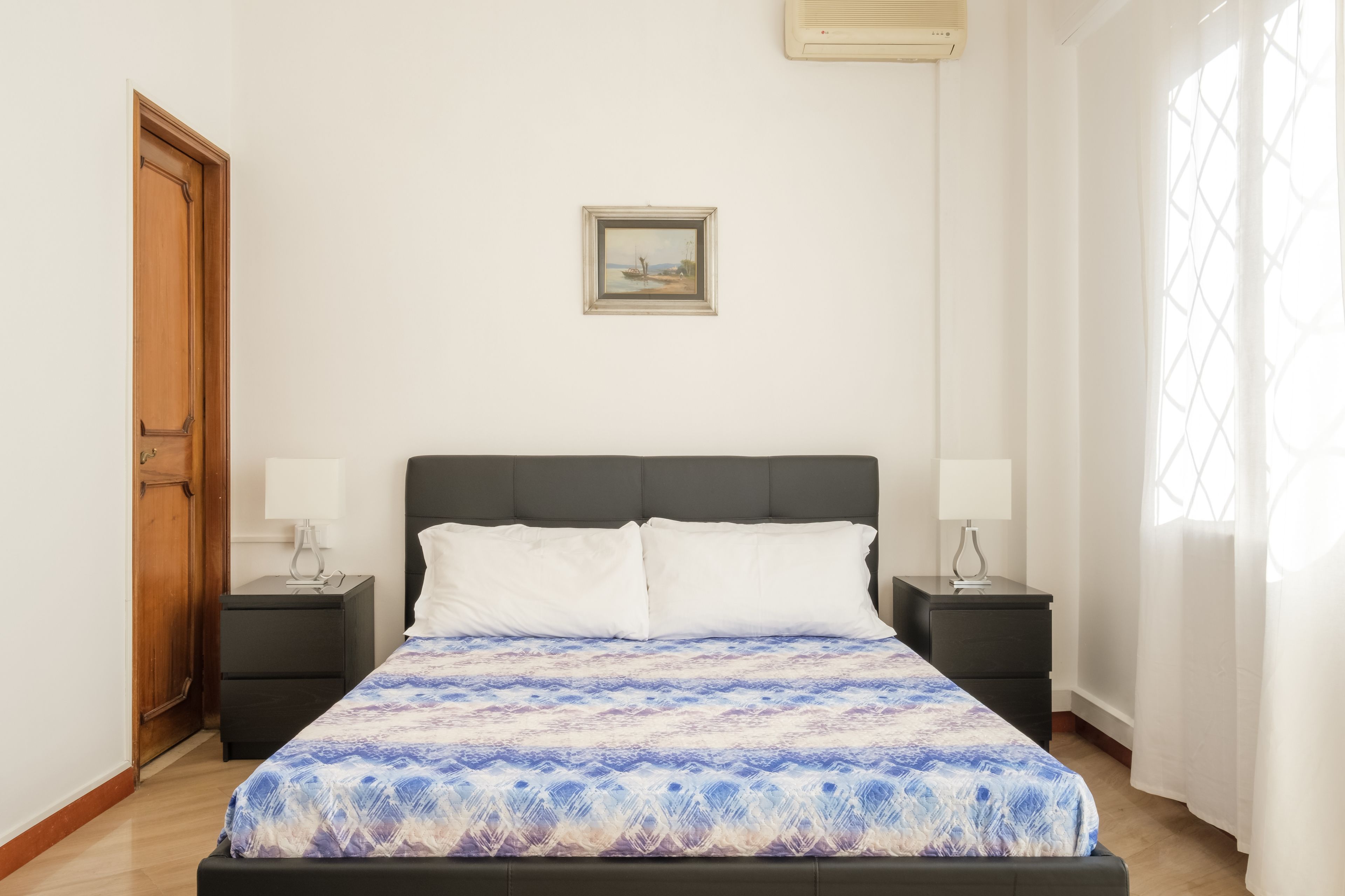 A bedroom with a bed, two bedside tables, a nightstand, a wooden door, a wall-mounted air conditioner, and a window with curtains.