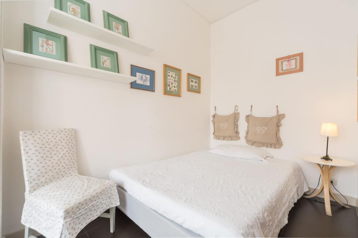 Een slaapkamer met een bed, een stoel, een klein tafeltje en wandplanken met ingekaderde foto's.