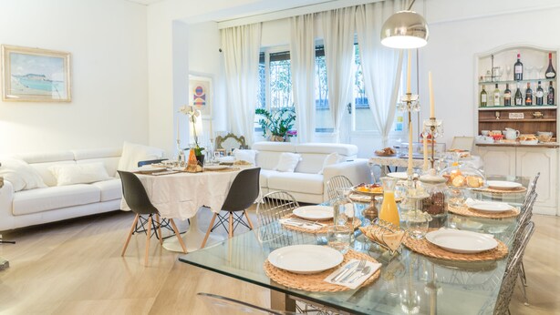 A dining area with a glass table set for six, surrounded by white sofas and a kitchen area with wine bottles.