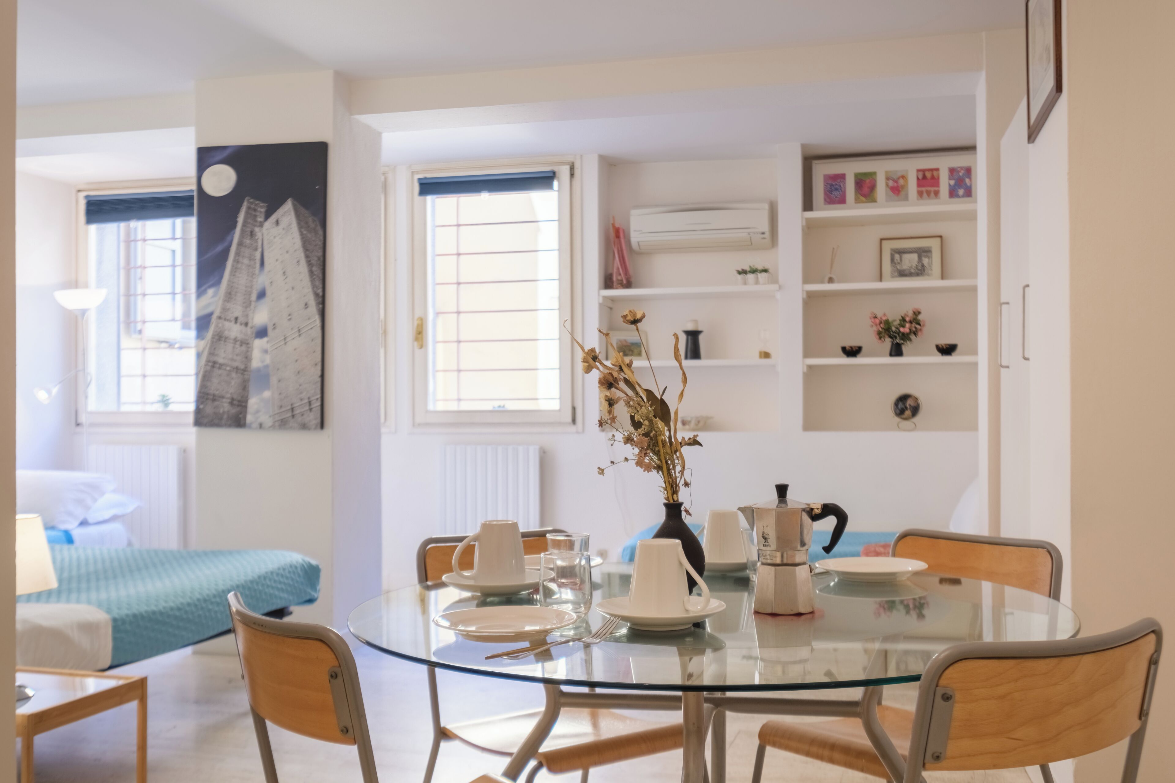 A modern dining area with a glass table, chairs, and a vase with dried flowers. There is a bed with a blue blanket in the background and a window with blinds.