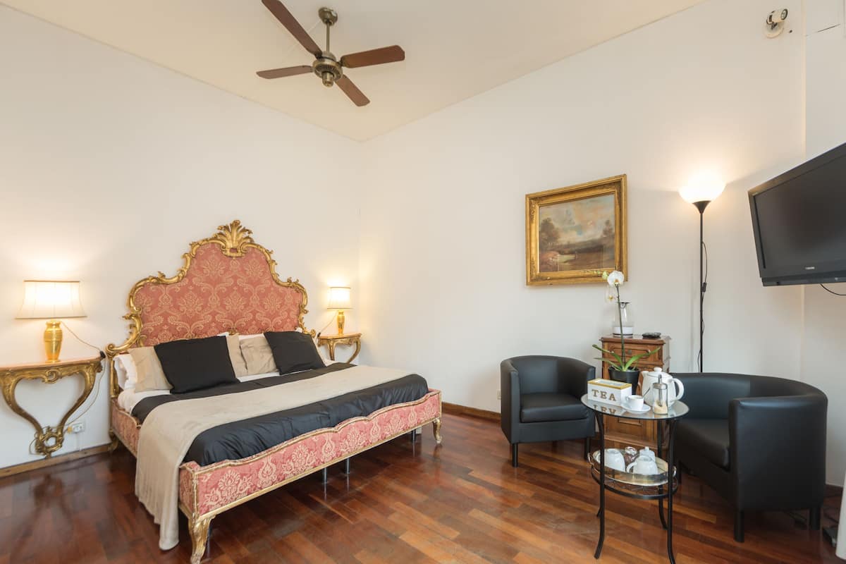 Een hotelkamer met een groot bed, twee nachtkastjes met lampen, een plafondventilator, een televisie, een inlijsting schilderij en een kleine tafel met theeservies.