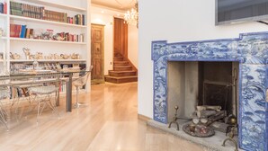A room with a bookshelf, a dining table with chairs, a fireplace with blue tiles, and a staircase.