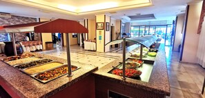 Zona de buffet con diversas opciones de comida, un panel de vidrio y vistas al vestíbulo del hotel.
