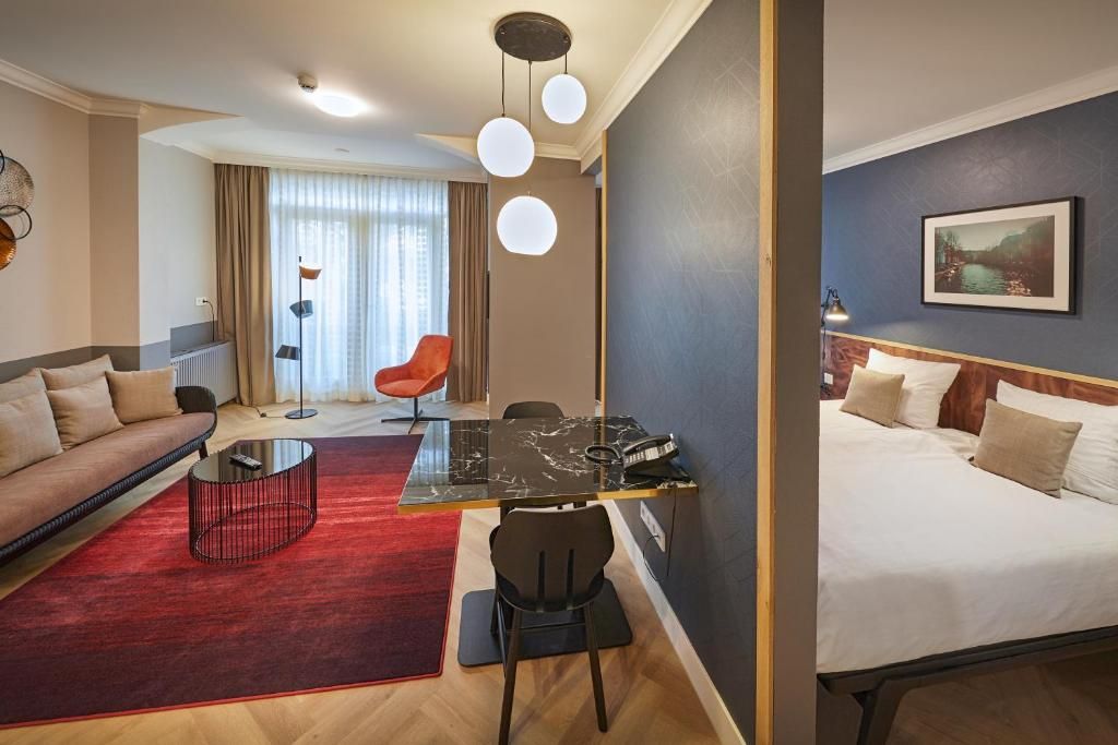 Habitación de hotel con cama, sofá, mesa de comedor y una silla. Hay una alfombra roja en el suelo y un cuadro en la pared.