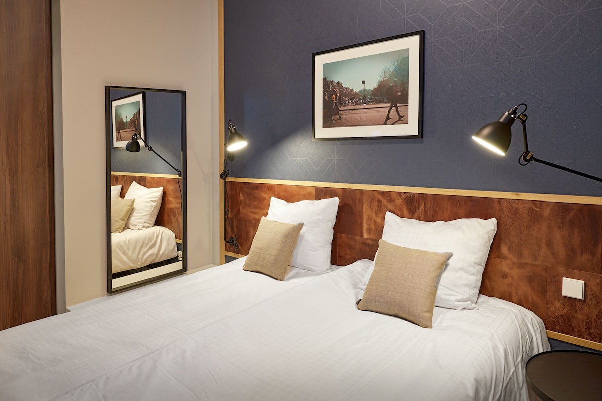 Un lit bien fait, avec du linge de lit blanc et des oreillers beige, une tête de lit en bois, un tableau encadré au mur et deux lampes de chevet.