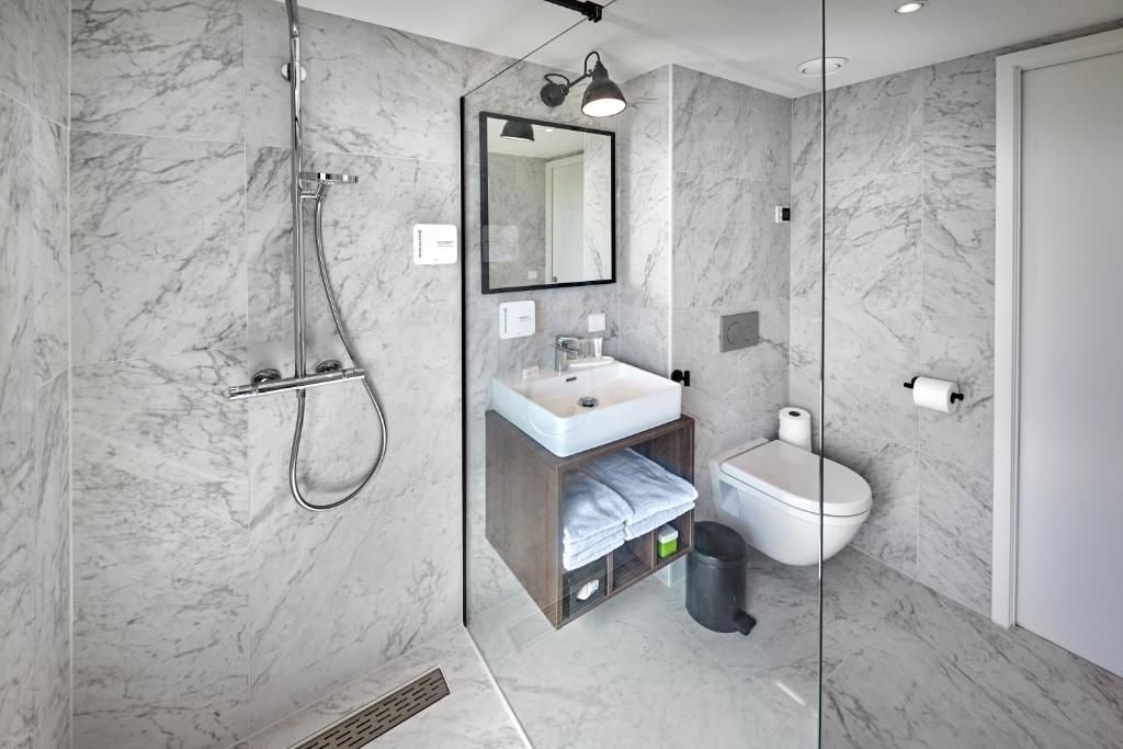 Un baño moderno con ducha de vidrio, pared con mosaicos de mármol, lavamanos con espejo y retrete.