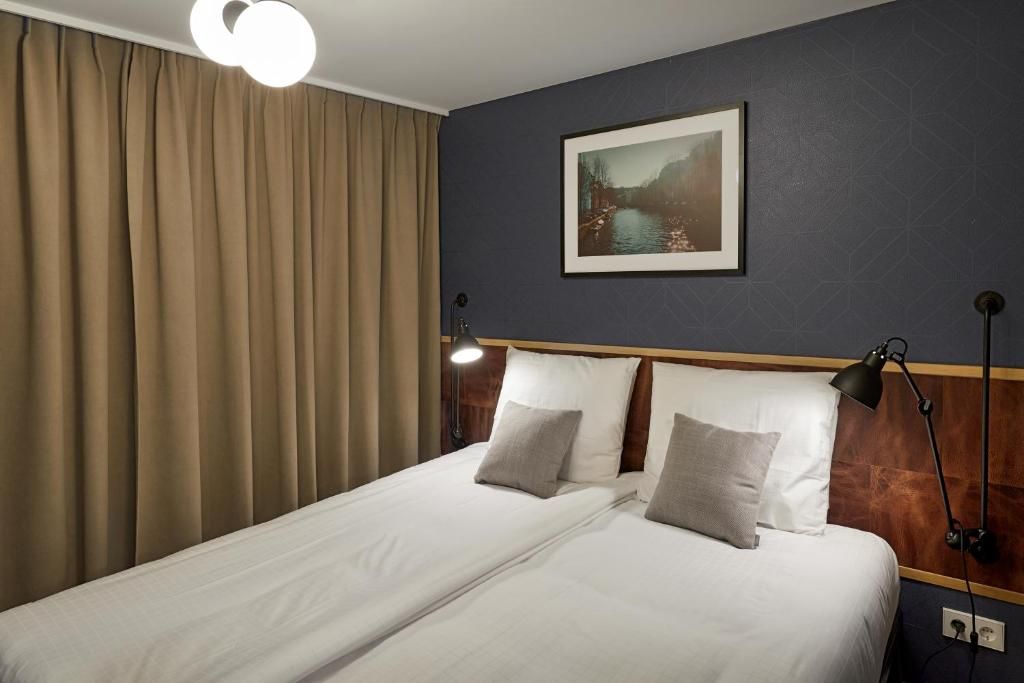 Habitación de hotel con cama, cortinas, una lámpara de pared y un cuadro enmarcado.