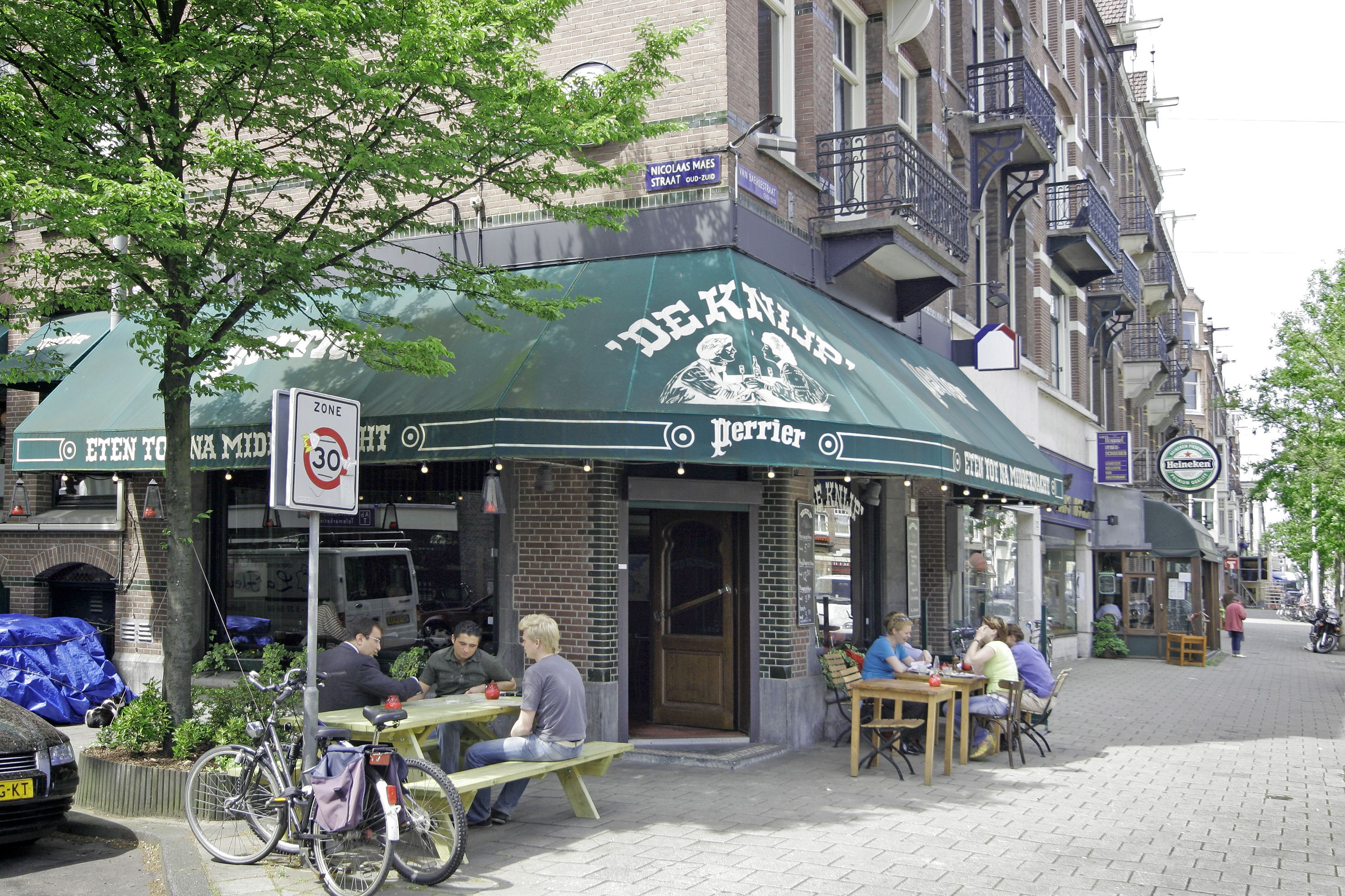 An outdoor café with tables and chairs, a bicycle parked nearby, and a building with a sign above the entrance.