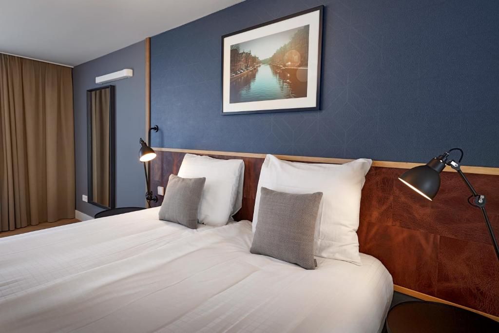 Ett hotellrum med en stor säng, två sänglampor, en inramad tavla på väggen och ett fönster med gardiner.