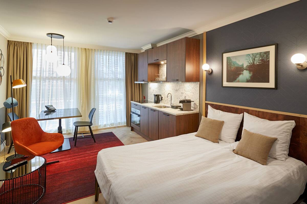 Une chambre d’hôtel comprenant un lit, un bureau, une chaise, un coin cuisine et une grande fenêtre avec des rideaux.