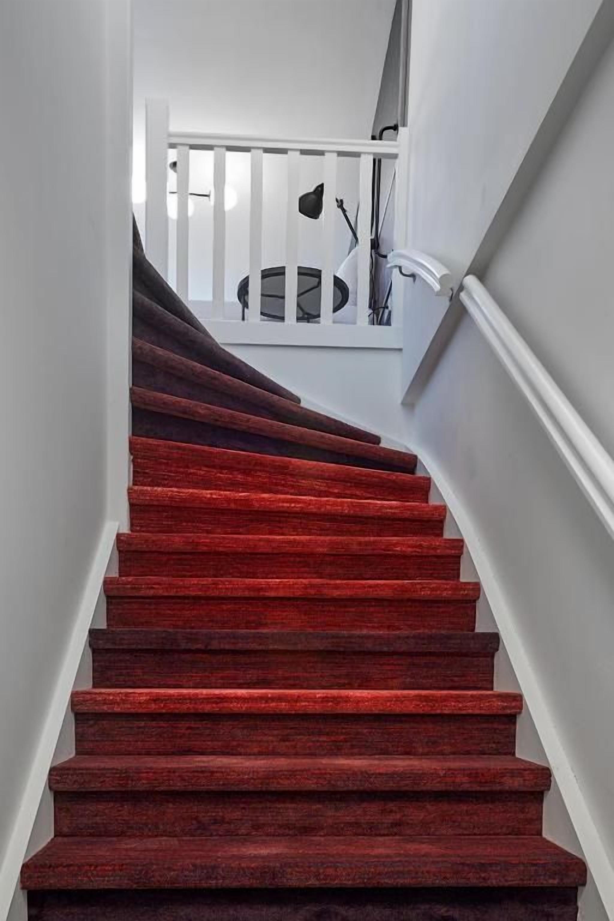Una escalera con alfombra roja, paredes blancas y barandilla blanca.