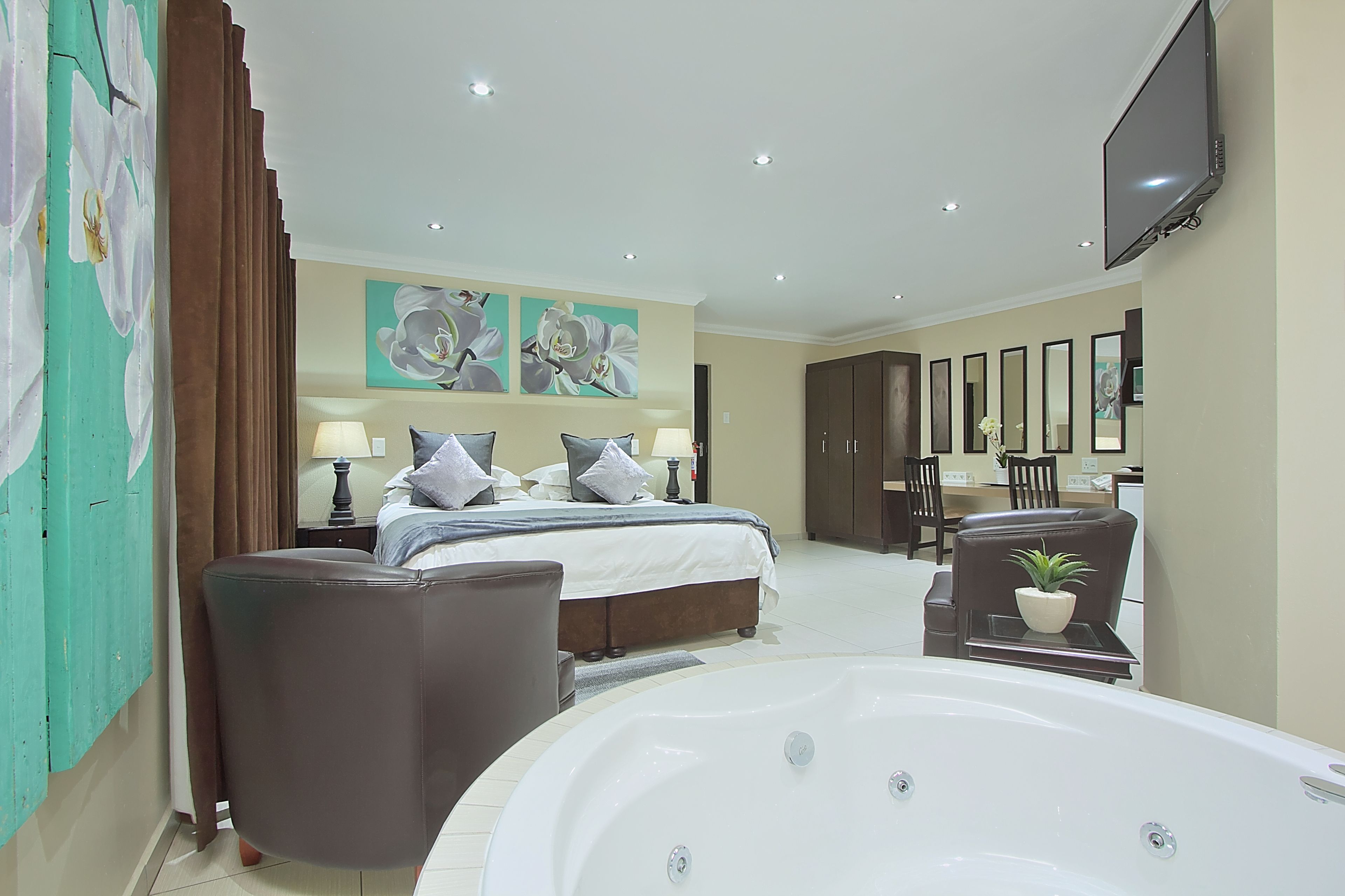 A modern hotel room with a large bed, a sitting area with a chair, a desk, a TV, and a large bathtub.