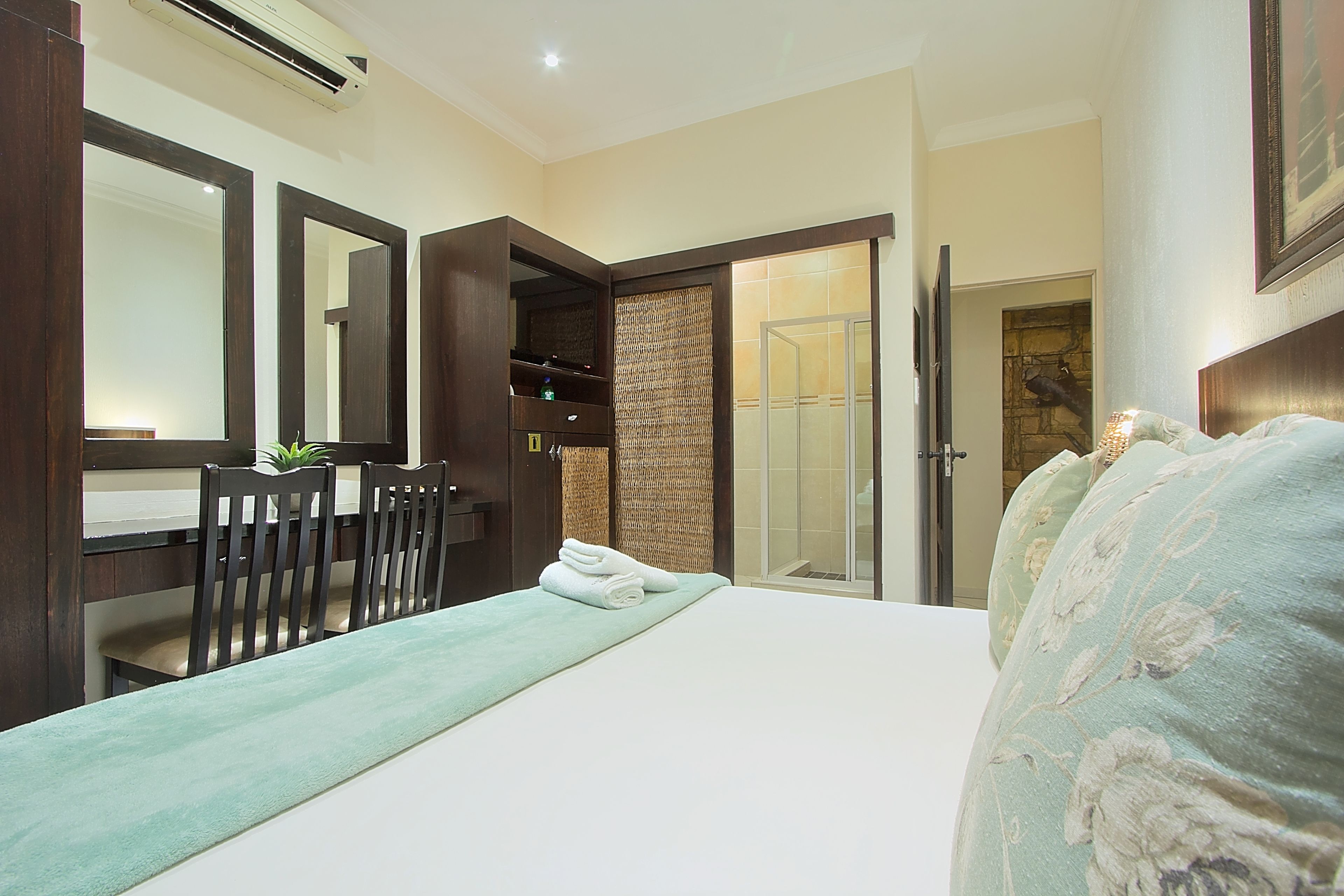 Habitación de hotel con cama, escritorio y sillas, un espejo grande y una puerta corredera que da a una zona de ducha.