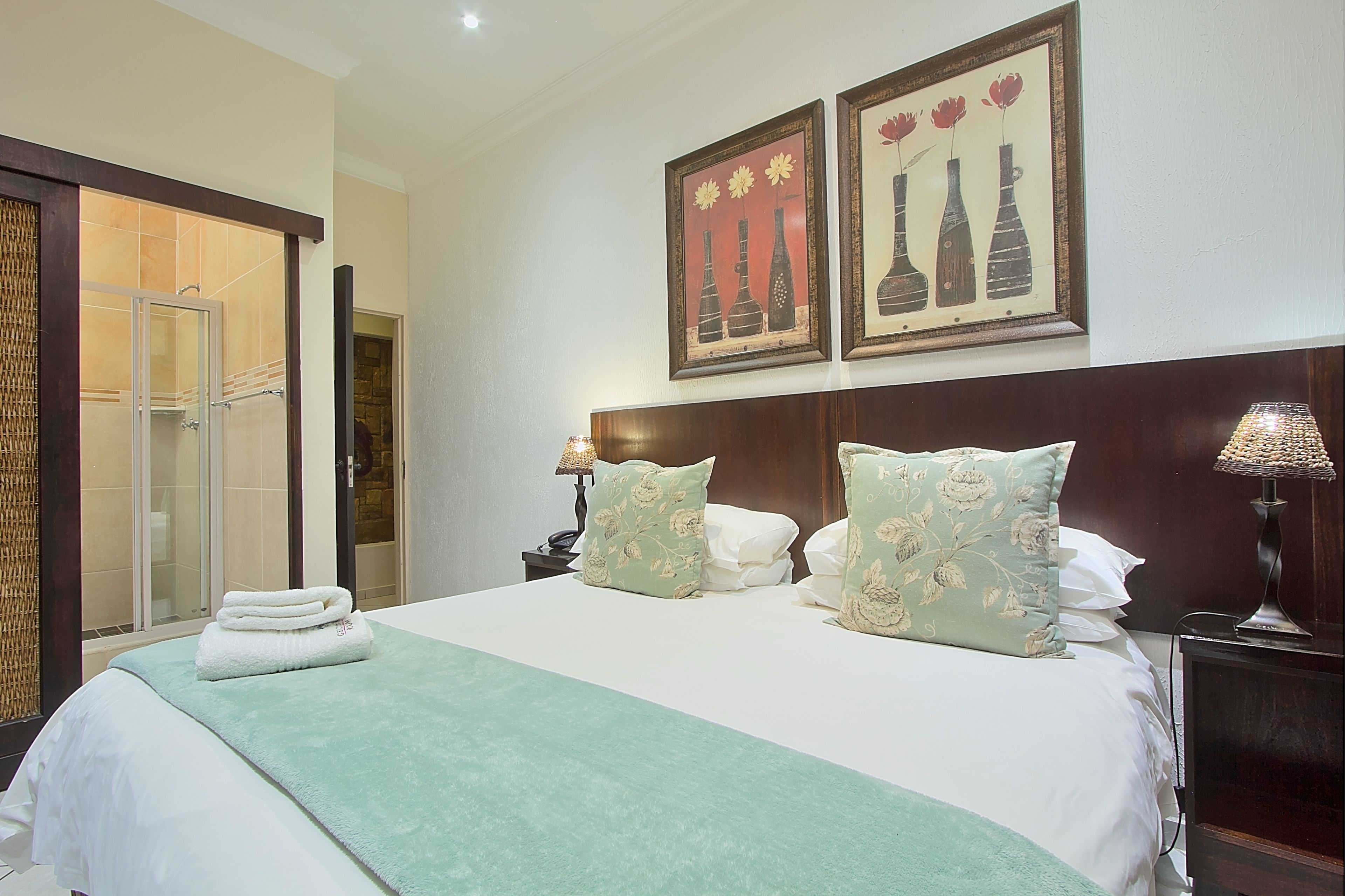 A hotel room with a bed, two nightstands, a lamp, a door leading to a shower area, and two framed artworks on the wall.