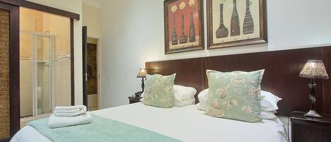 A hotel room with a bed, two nightstands, a lamp, a door leading to a shower area, and two framed artworks on the wall.