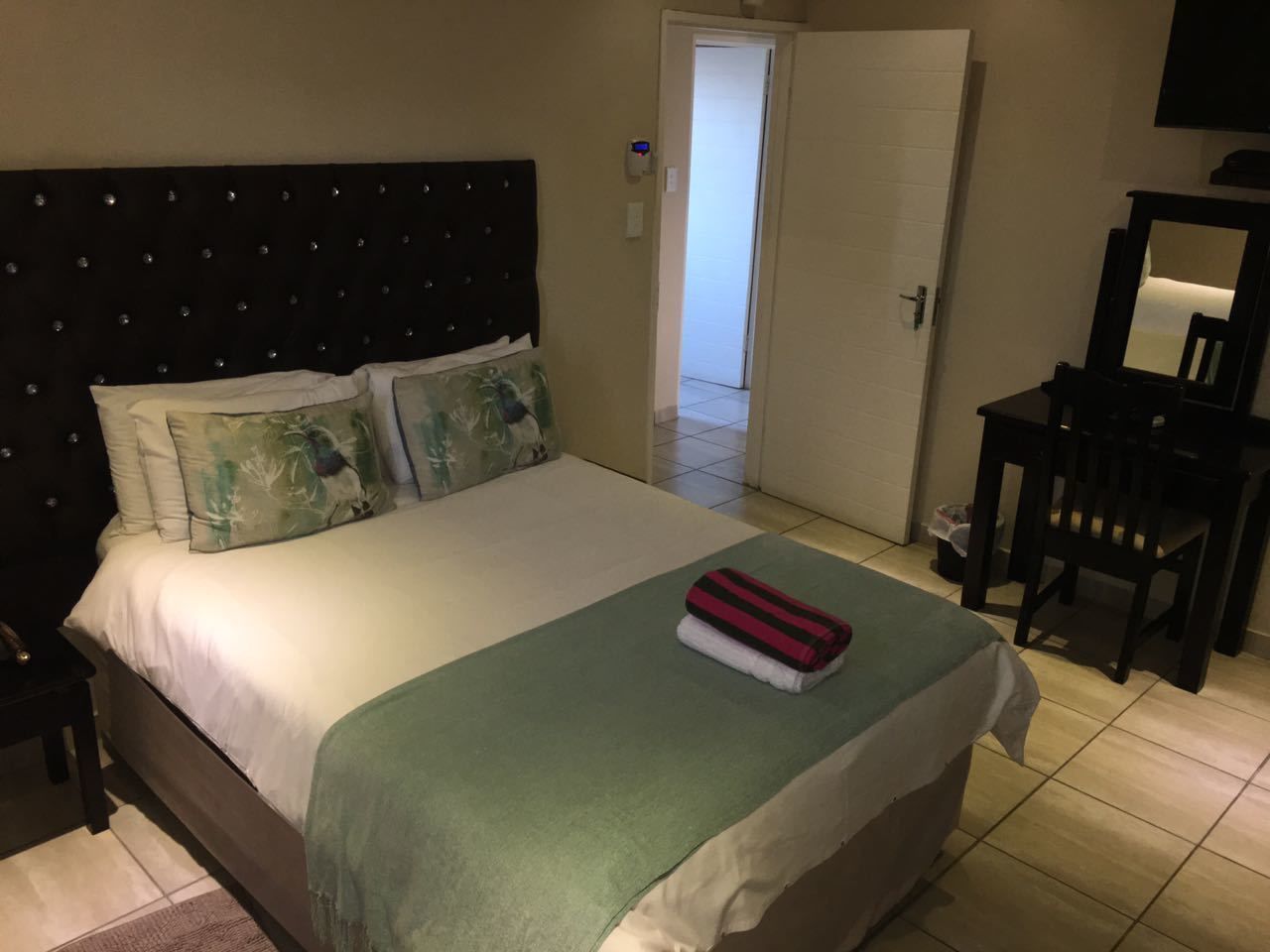 A neatly made bed with a green bedspread and pillows, a bedside table with a lamp, and a chair in a room with a tiled floor and a door leading to another room.