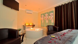 Ein Hotelzimmer mit einer Couch, einer runden Badewanne, einem Esstisch mit Kerzen und einem Bett mit Rosenblättern.