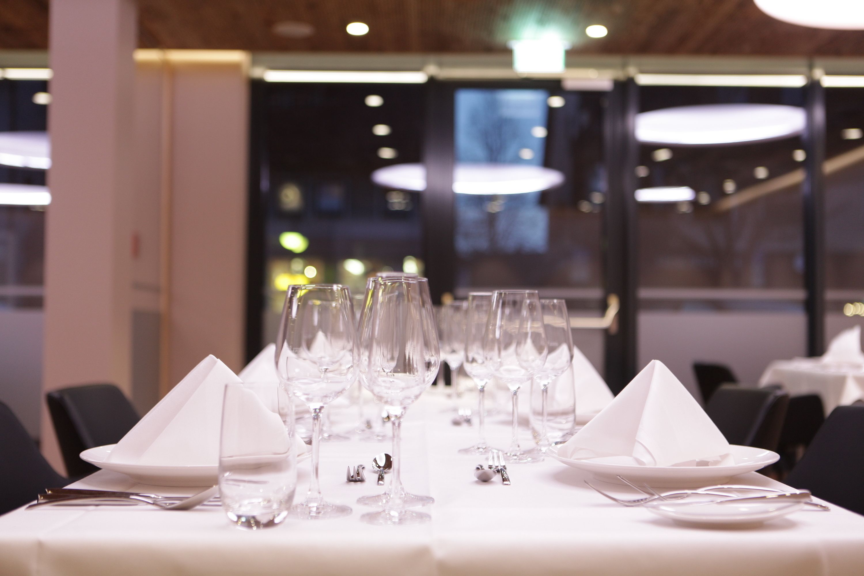 Mesa de comedor dispuesta con mantel blanco, vasos y cubiertos, en un restaurante moderno con vistas a la ciudad de noche.