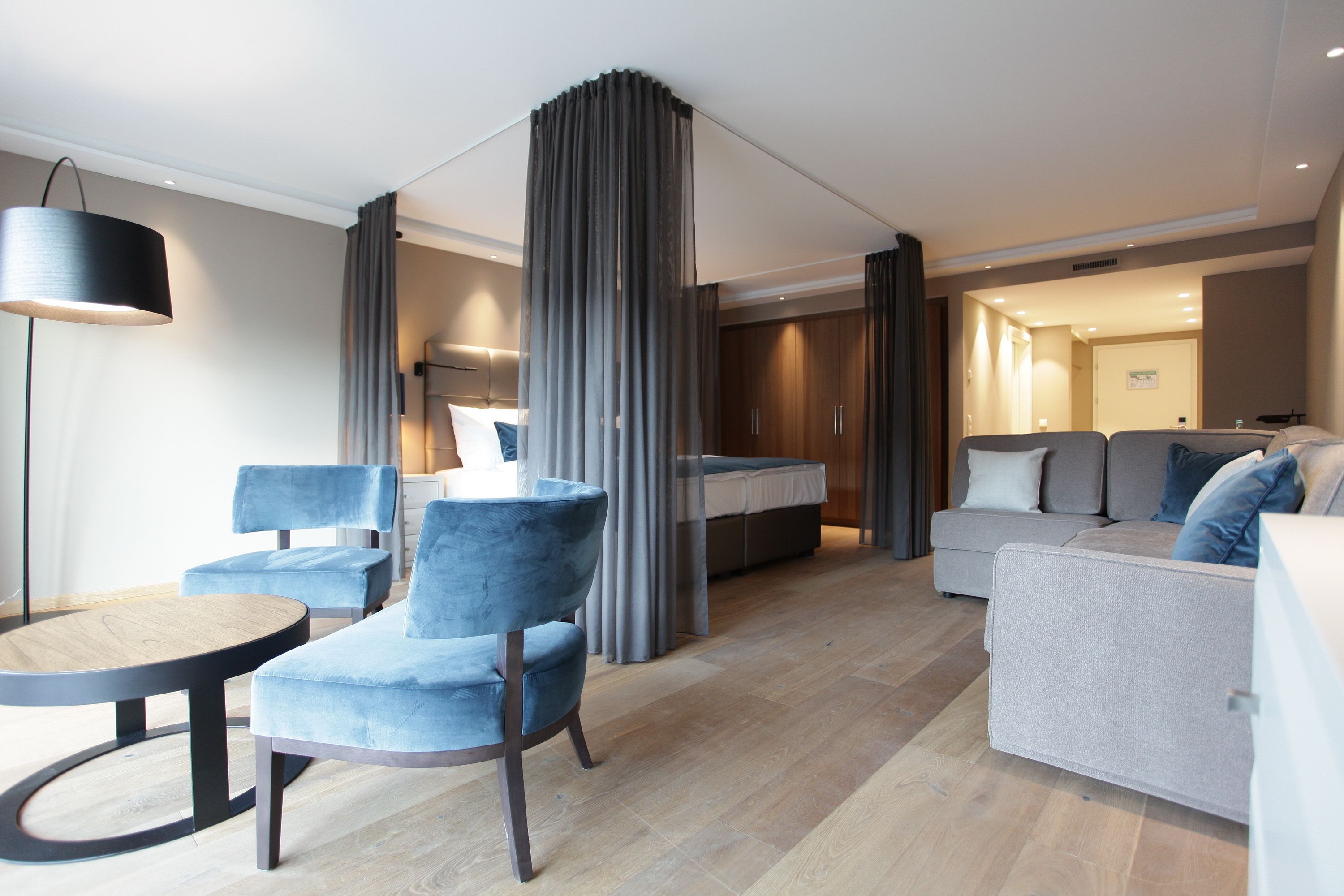 A modern hotel room with a sofa, two armchairs, a round table, a bed, and a large window.