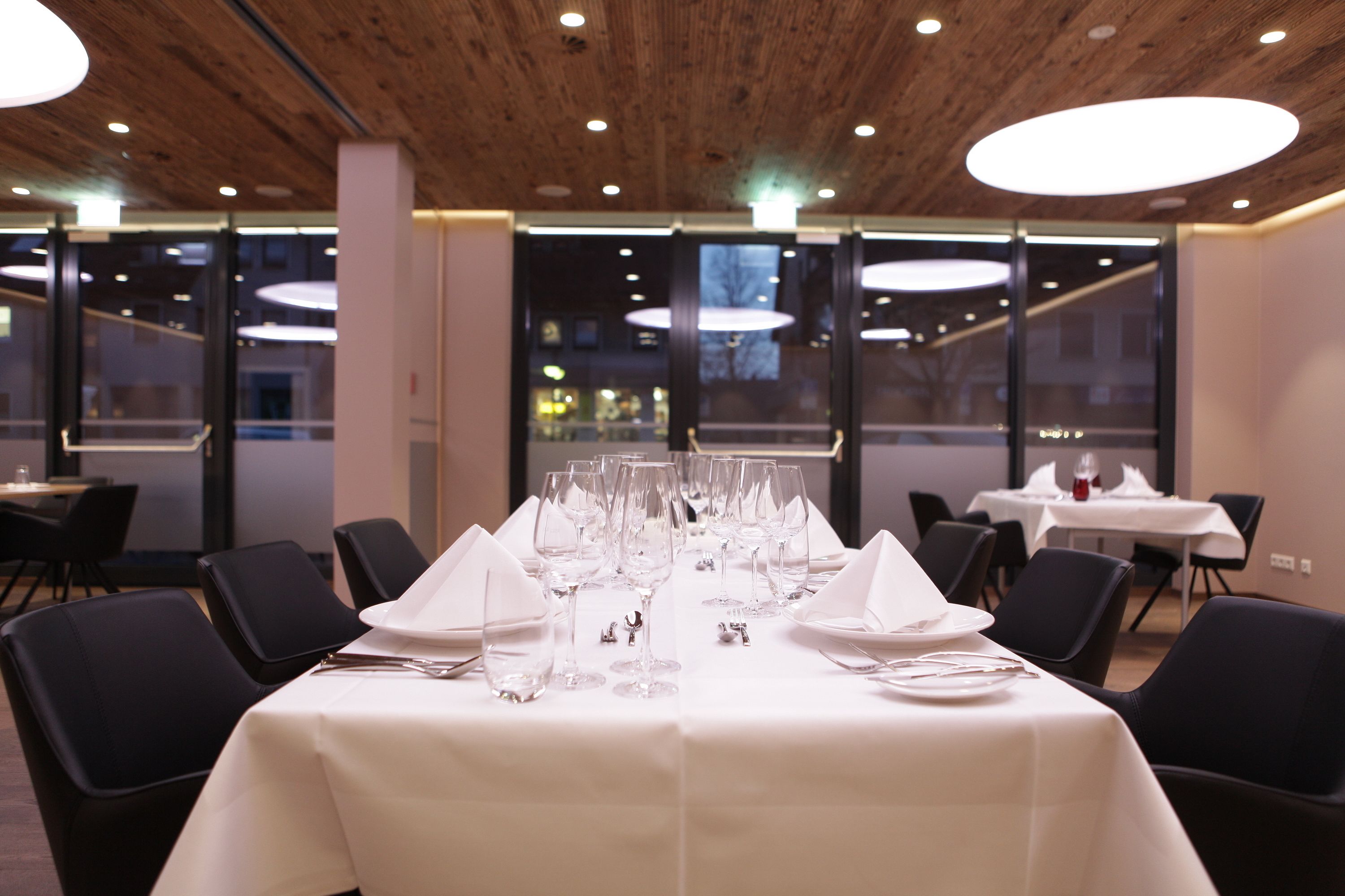 A dining area with tables set for a meal, featuring white tablecloths, black chairs, and a modern ceiling design.