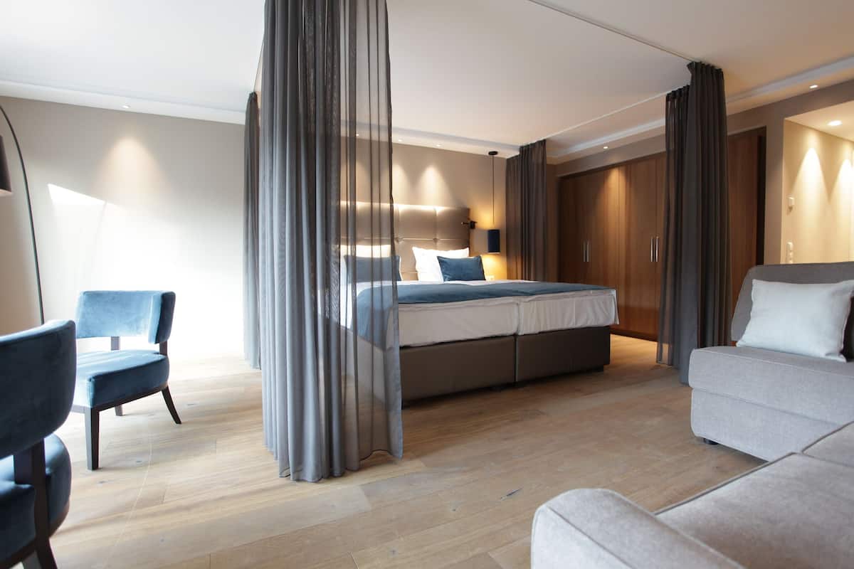 A modern hotel room with a large bed, a sofa, a chair, and a wardrobe.