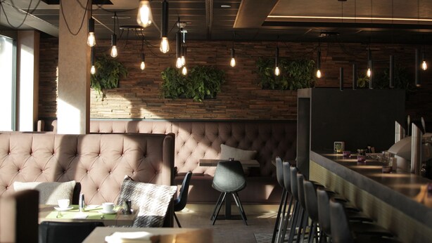 A modern interior with a tufted sofa, dining area, and bar with hanging lights and plants. - Hotel Aurelia (Aldingen)