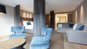 A modern hotel room with a bed, a grey sofa, two blue armchairs, and a wooden table. - Hotel Aurelia (Aldingen)