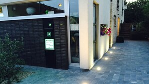 A building with a black exterior wall, a sign indicating 'Kookhai Thai Away Cooking Lessons', and a paved entrance area with a small bush. - B&B Kookhai (Kluisbergen)