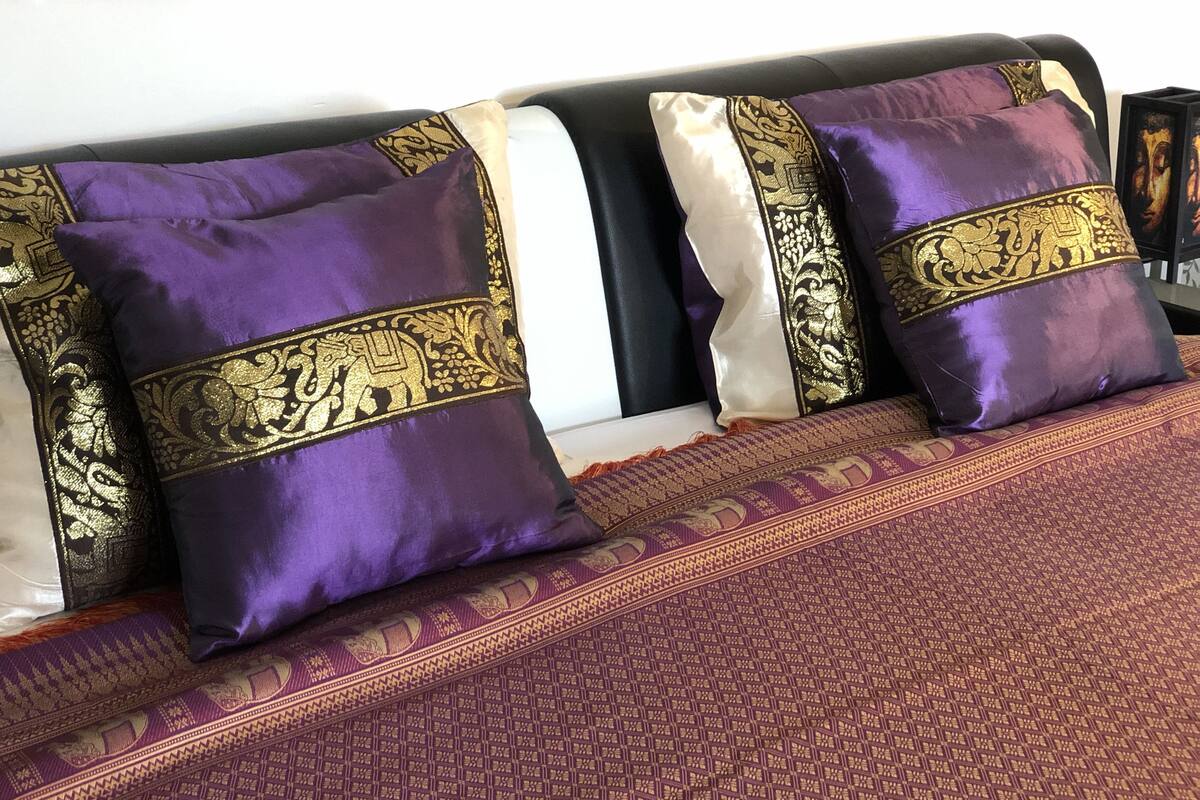 Uma cama com almofadas roxas e douradas, um edredom estampado e uma almofada branca.