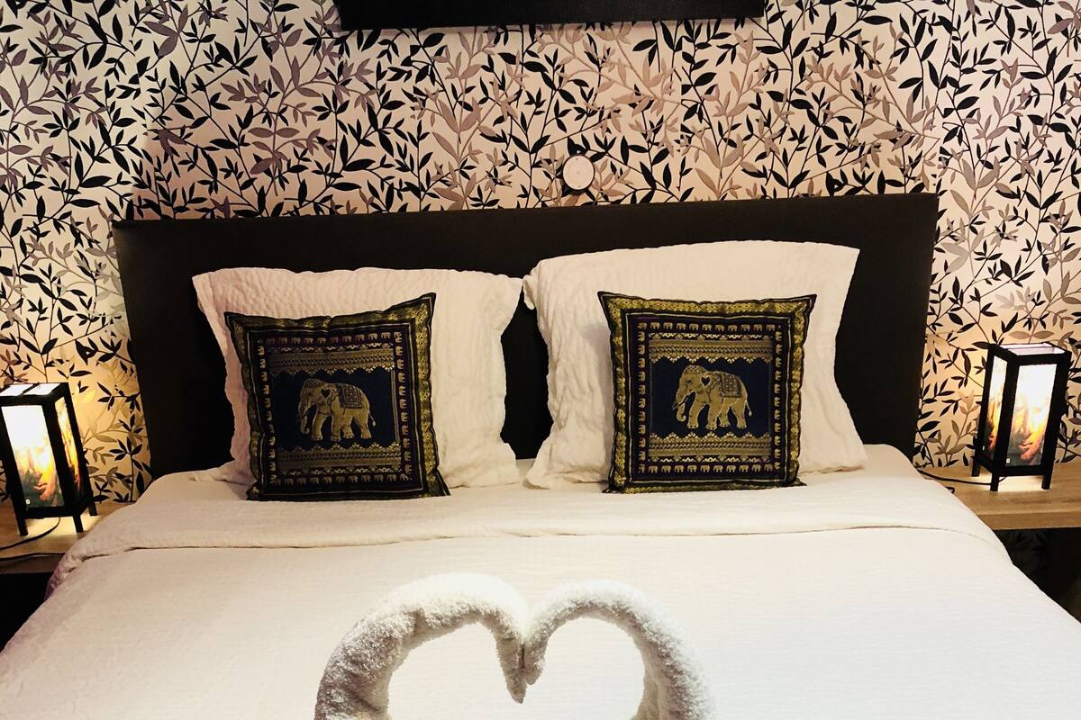 Cama bem arrumada com arranjo de toalha em formato de coração e duas flores cor-de-rosa.