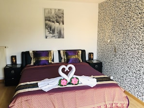 Una cama bien hecha con una toalla dispuesta en forma de corazón, almohadas moradas y doradas, y una pared con diseño.