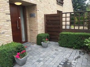 Un edificio de ladrillo con puerta y portón de madera, plantas en macetas y un área de entrada pavimentada.