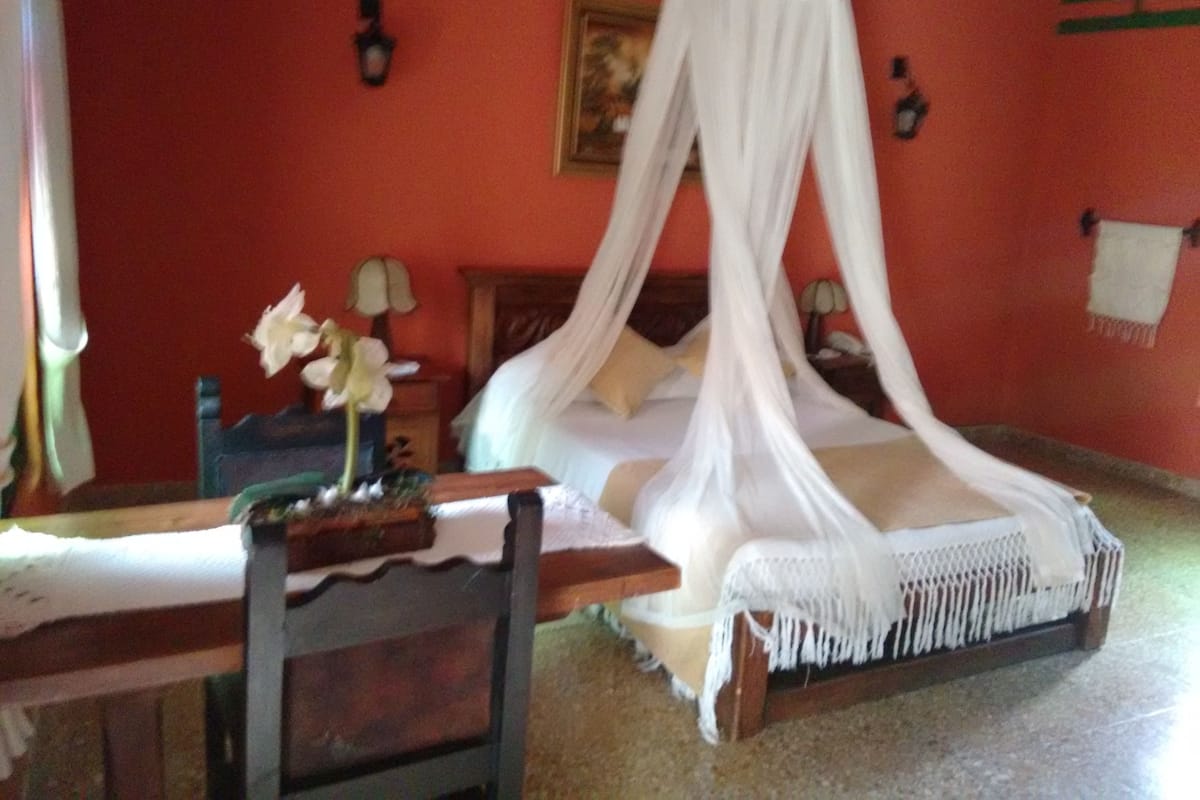 A room with a four-poster bed, a wooden nightstand, a table with a vase of flowers, and a wall-mounted lamp.
